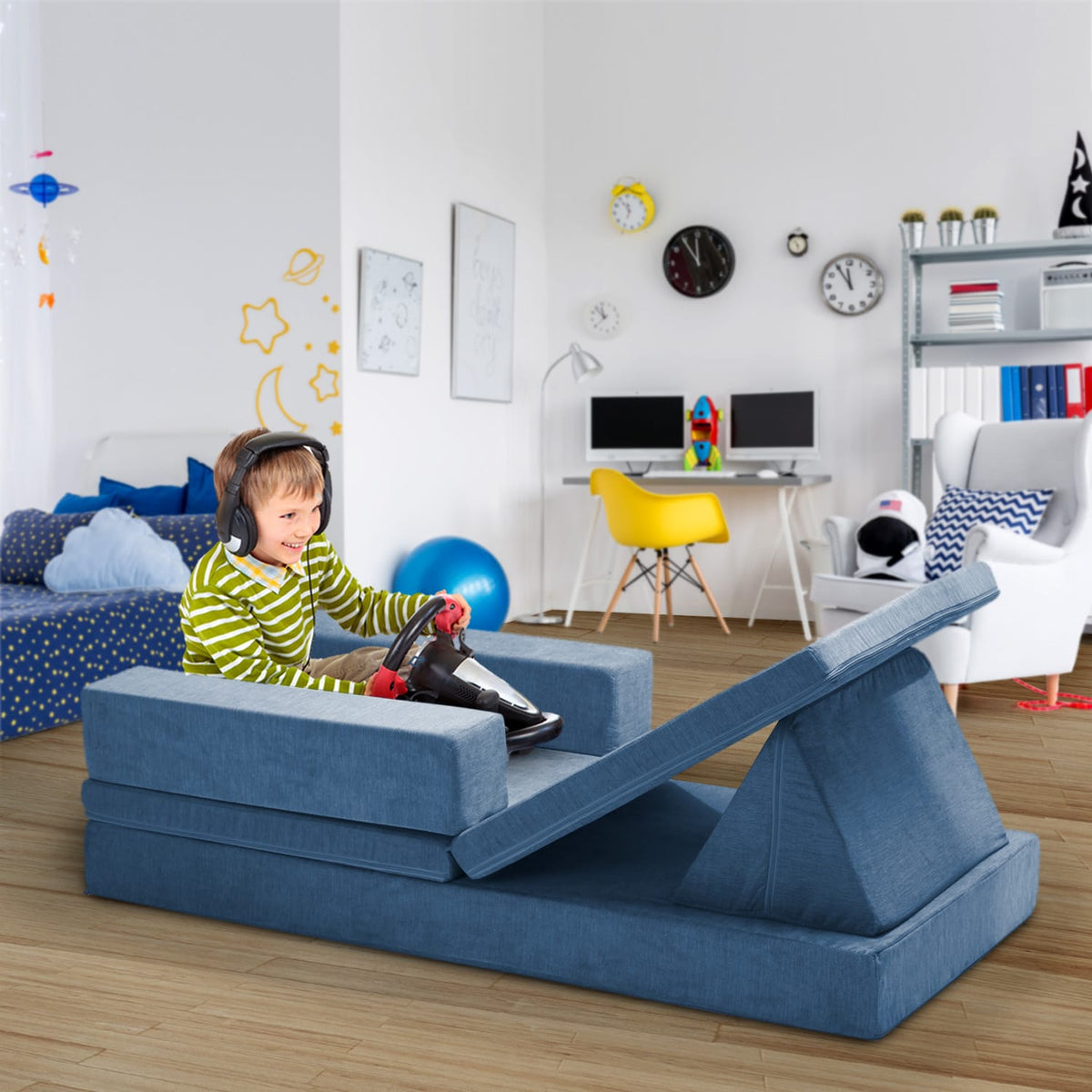 A child wearing headphones sits on a blue modular FoamOh foam play couch in a child's room, pushing the couch forward with a toy car. The room features a desk, chair, and shelves in the background.