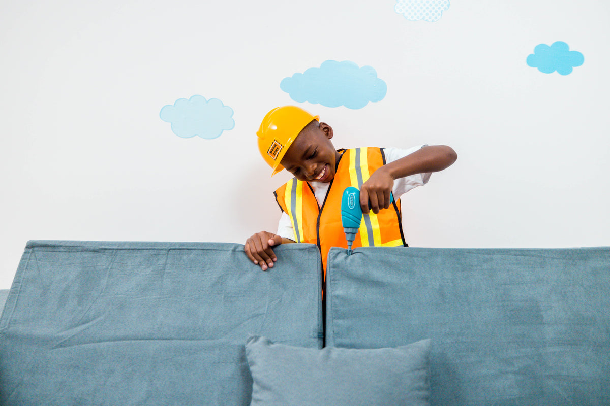 A young boy dressed as a construction worker, wearing a yellow hard hat and an orange safety vest, playfully peering over the back of a blue fabric kids sofa against a white wall with light blue cloud decals.