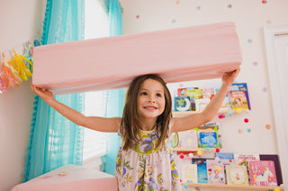 A young girl with long brown hair smiles while holding a large, rectangular pink foam block above her head in a brightly lit children's room with turquoise curtains and colorful bookshelves.FoamOh castle kids sofa