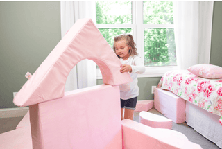 A young girl stands in a child's bedroom playing with a large pink foam house structure made of soft play blocks. The room has a window with white curtains, a bed with pink floral bedding, and additional pink foam play pieces on the floor.