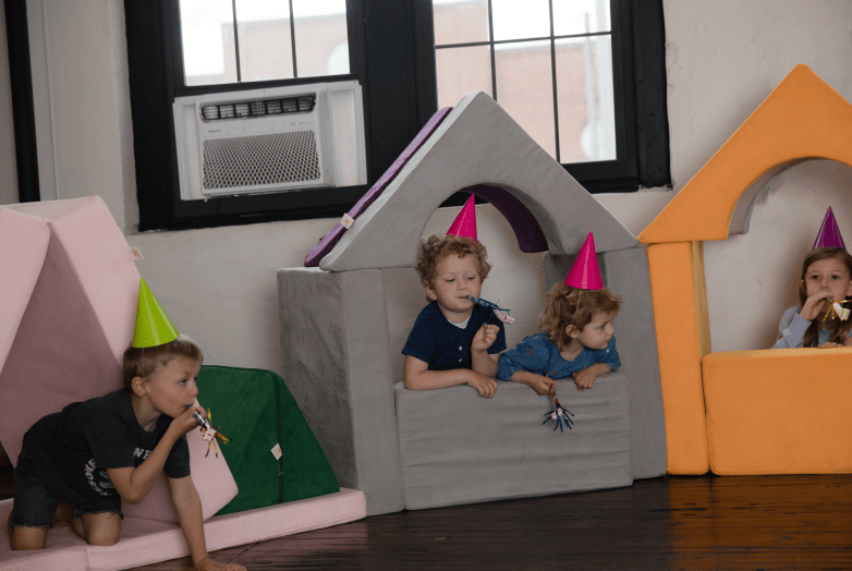 Three children wearing pink party hats play inside colorful FoamOh blocks - foam playhouses in a classroom or playroom setting.