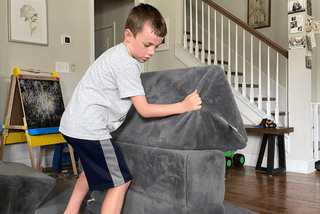 A young boy in a gray t-shirt and navy shorts pushes a dark gray recliner chair in a living room with wooden floors and a staircase in the background.