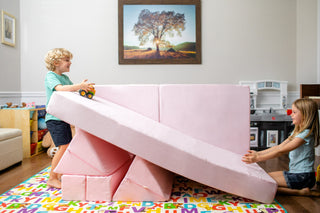 Two children playing with a FoamOh large pink foam wedge ramp in a child's playroom. One child is standing on the left side holding a small yellow toy car, and the other is kneeling on the right side pushing the ramp.