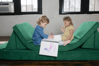Two young children sitting on a green FoamOh castle sofa, drawing in a spiral notebook on the floor between them in a bright room with large windows.