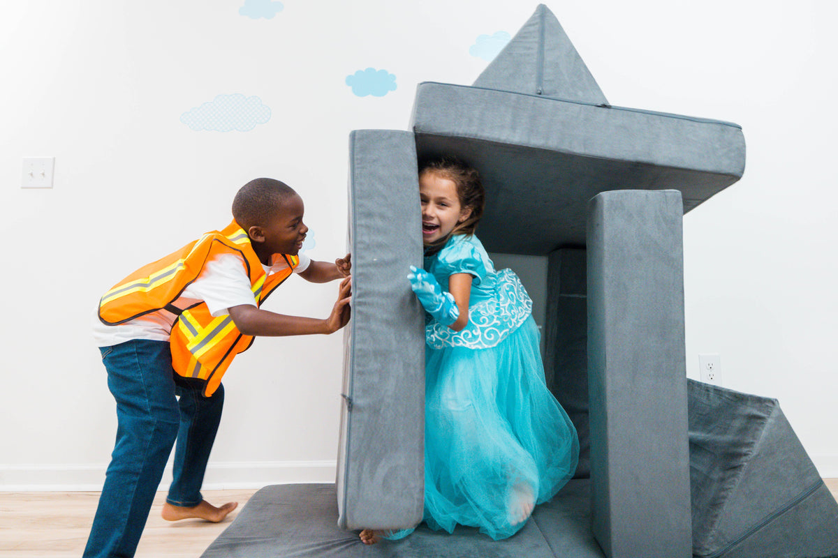 A young boy in a high-visibility safety vest and jeans pushes a girl in a blue princess dress inside a large gray foam play castle in a child-friendly room.