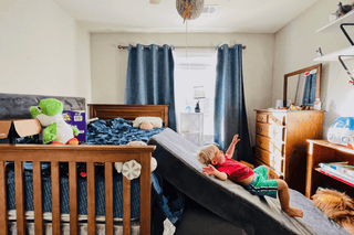 A child in a red shirt and green shorts lies on a mattress stacked on top of a wooden bed frame in a child's bedroom. The room contains a wooden dresser, a window with blue curtains, and various toys and furniture.