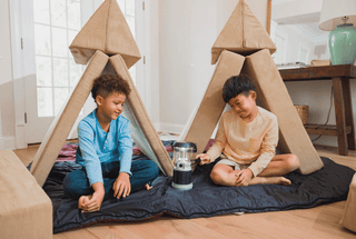 Two children sitting inside large cardboard teepee playhouses on a dark sleeping bag in a bright room, with a small lantern between them.