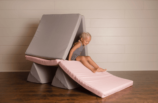 A young child sits between two padded foam blocks in a gym-like setting with a light wood paneled wall and dark wood floor. The Foamoh building blocks are upholstered in light gray and pale pink fabric.
