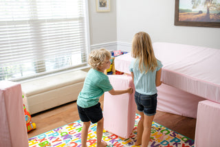 Two children pushing a FoamOh pink storage ottoman in a brightly lit children's room with a bed, colorful alphabet rug, and toys.  kids play couch