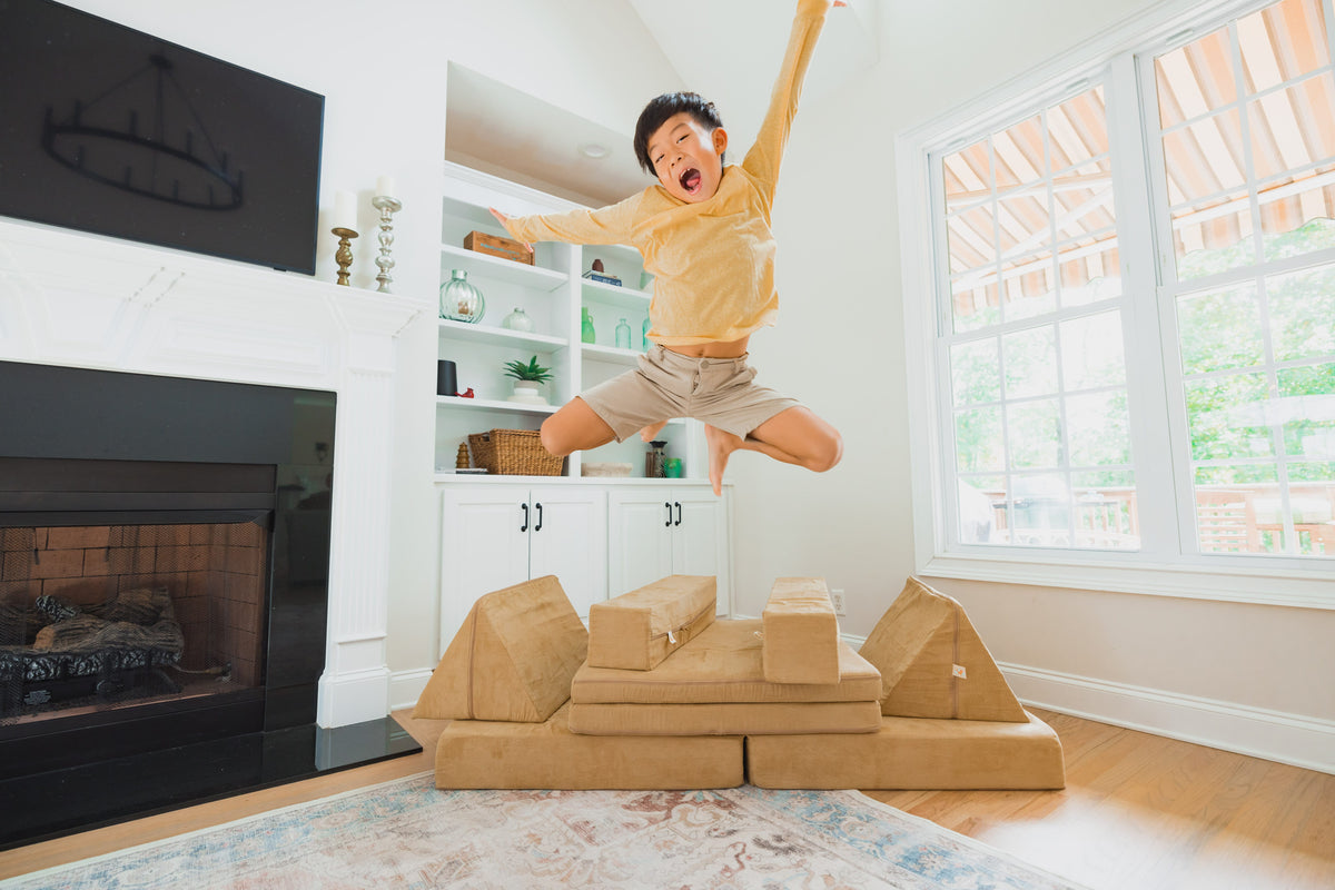 A young boy wearing a yellow long-sleeve shirt and khaki shorts jumps excitedly over a stack of FoamOh memory foam building blocks from a kids play couch in a bright, modern living room with a fireplace and large windows.