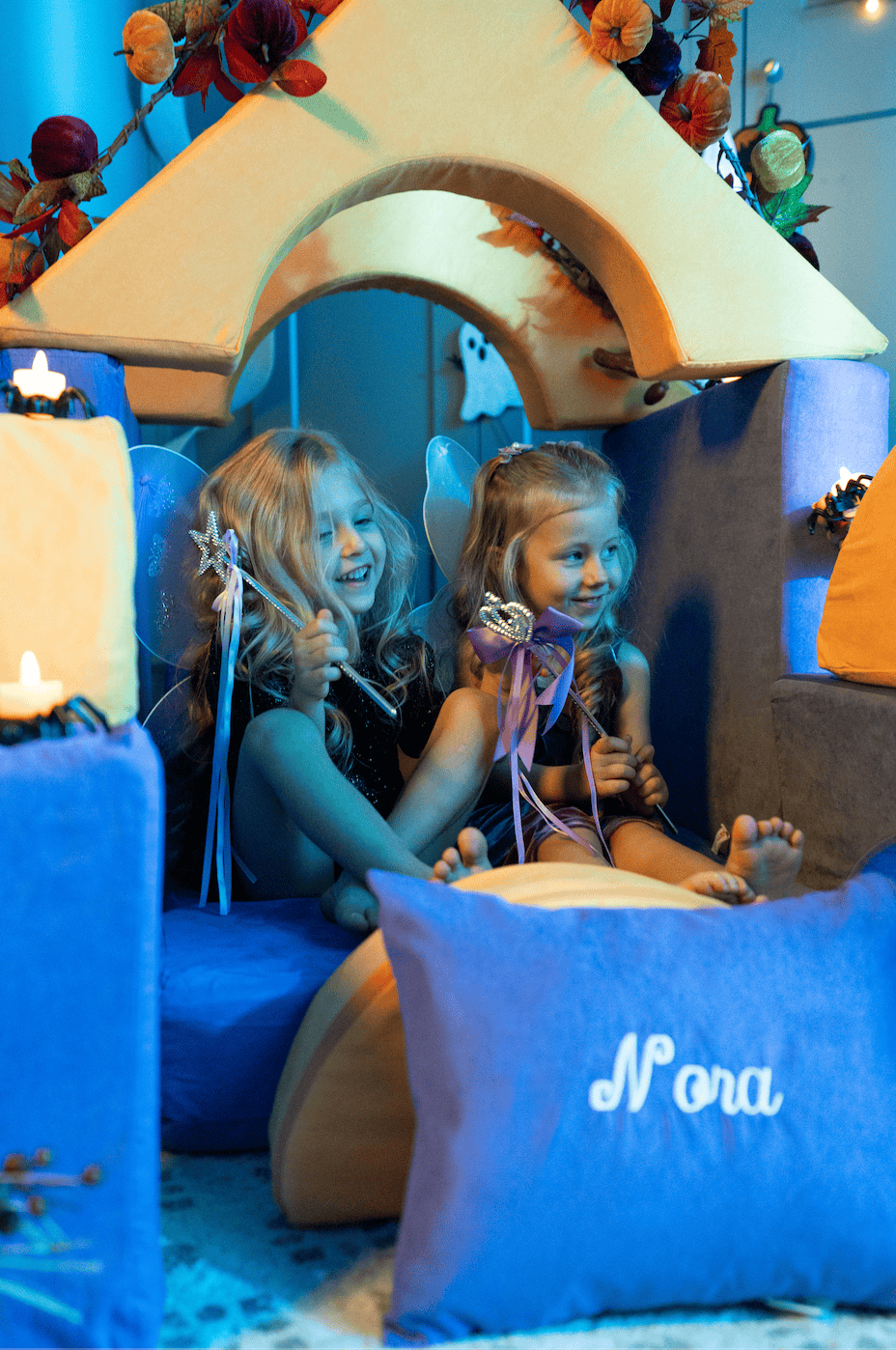 Two smiling girls in fairy wings and tiaras sit together on a FoamOh foam throne with a personalized “Nora” pillow.