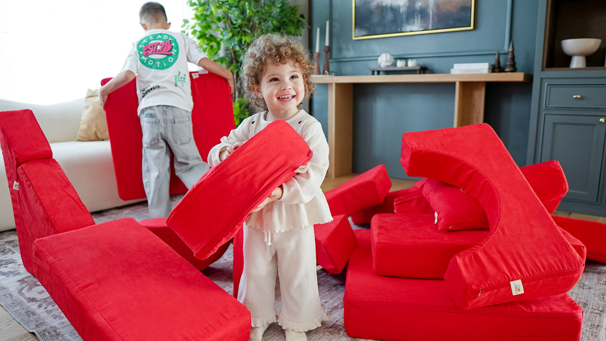 Smiling toddler carrying a red FoamOh kids play couch piece while her brother stacks more pieces in the living room