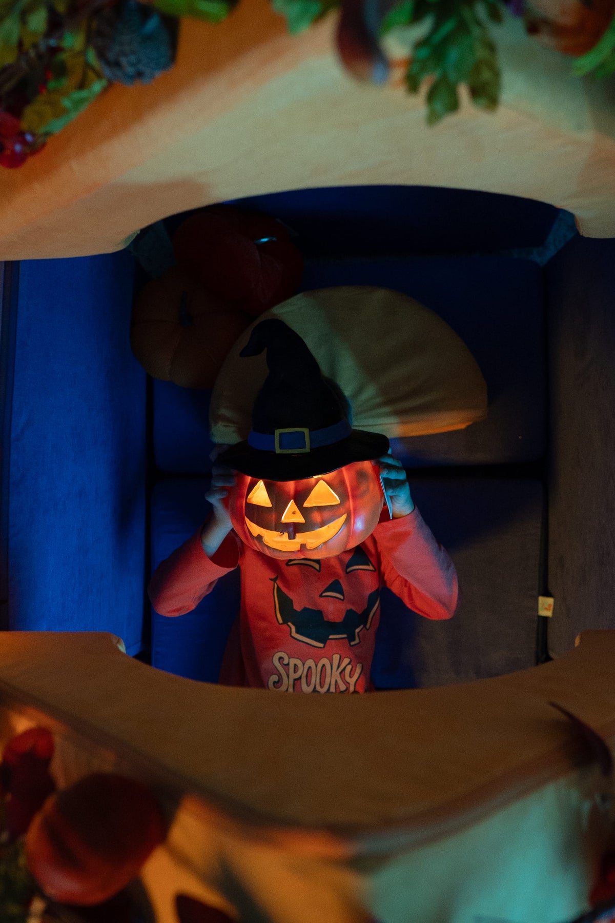 Child wearing a glowing jack-o’-lantern mask and witch hat peers through the window of a FoamOh foam fort