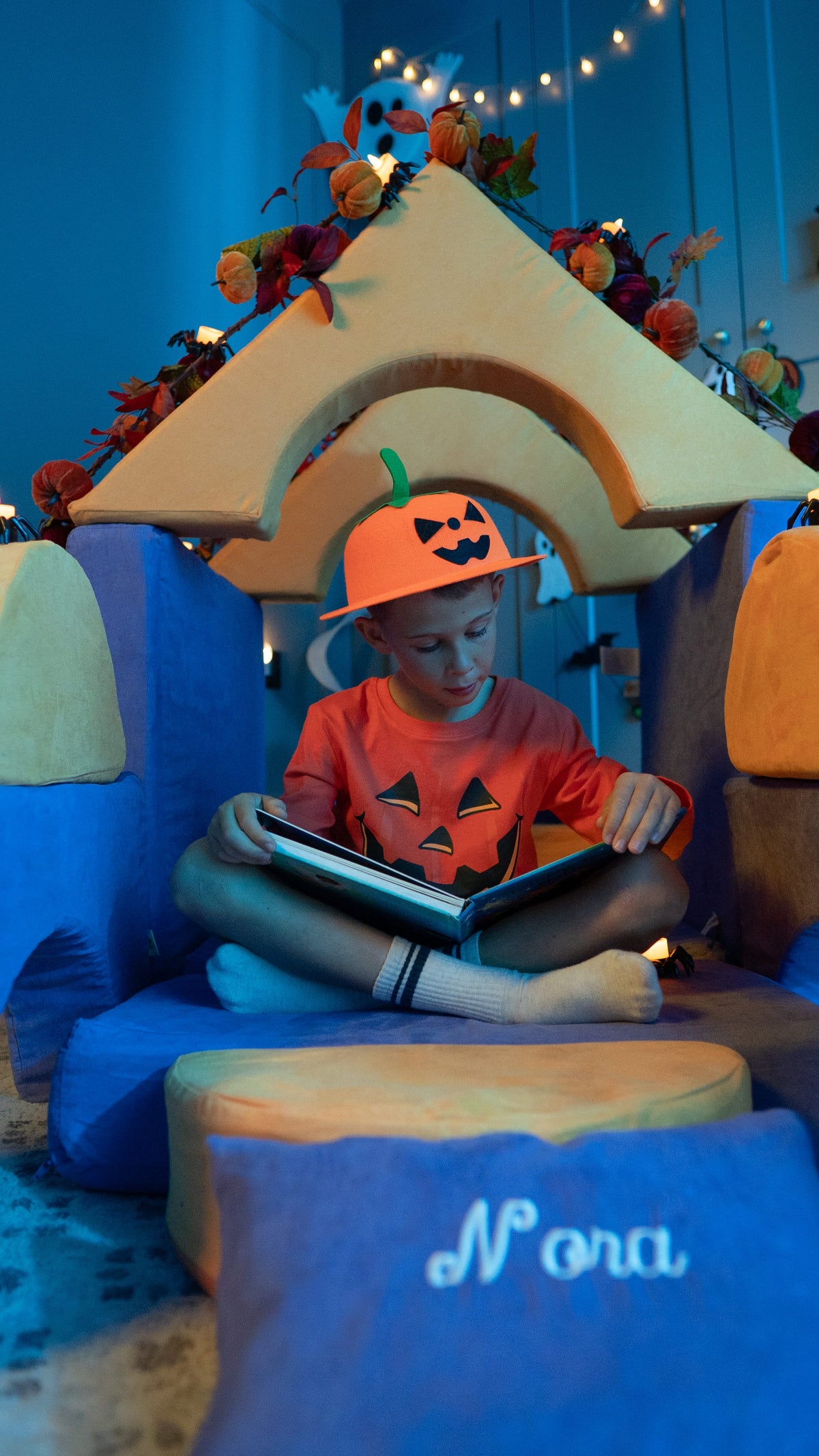 Boy in a jack-o’-lantern shirt and pumpkin hat sits cross-legged reading a book inside a cozy FoamOh foam fort with an arched roof.