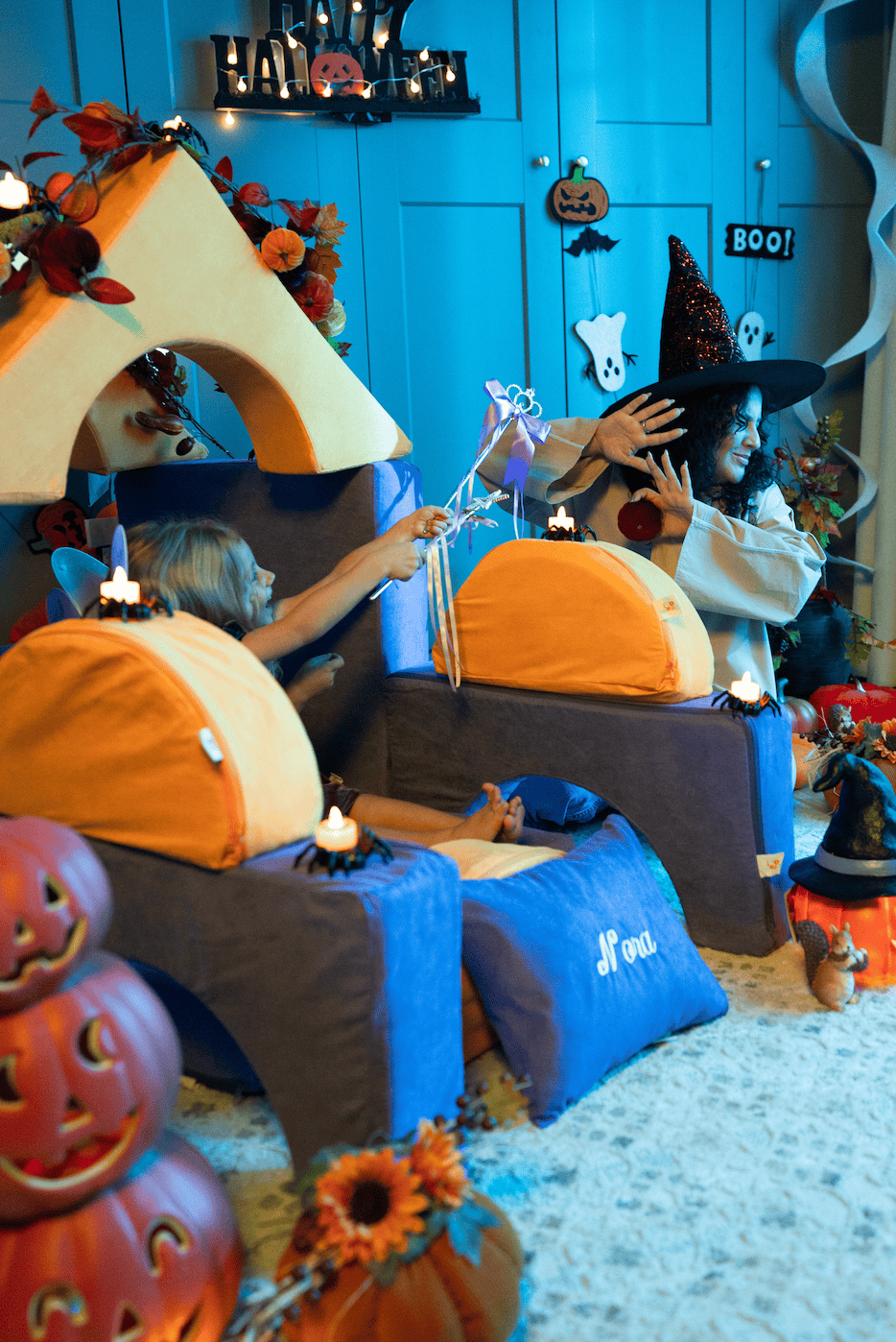 Girl in a witch hat waves a wand while another child peeks out from behind a FoamOh foam fort decorated with jack-o’-lanterns and candles.